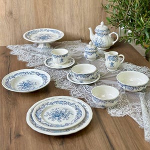 Può includere: Un servizio da tè in porcellana bianca con un motivo floreale blu. Il set comprende una teiera, tazze, piattini, ciotole, piatti, una zuccheriera, una lattiera e un alzatina per torte. Gli articoli sono disposti su una tovaglia di pizzo.