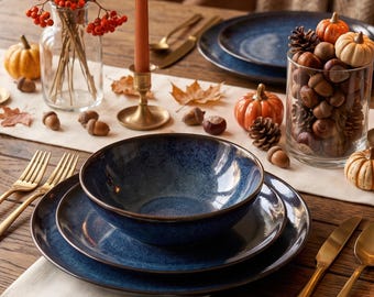 May include: A dining table setting featuring dark blue ceramic plates and bowls, gold-toned flatware, and orange taper candles in brass holders. A glass vase holds dried grasses and red berries, while a glass jar contains acorns and mini pumpkins.