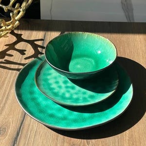 Pode incluir: Um conjunto de três peças de louça de cerâmica em tons de verde. O conjunto inclui uma tigela e dois pratos, todos com um esmalte rachado texturizado e bordas prateadas. Os pratos estão dispostos sobre uma superfície de madeira, com uma peça decorativa dourada no fundo.