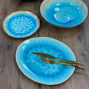 Op de afbeelding: Een set van vier blauwe en gouden keramische serviesdelen, waaronder een grote ovale schaal, een kleinere ronde plaat, een kom en een kleinere kom. Het servies heeft een blauwe glazuur met een subtiel marmer effect en een gouden rand.