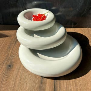 Könnte beinhalten: Ein Satz von drei gestapelten, cremeweißen Keramikschalen. Die obere Schale enthält eine Gruppe leuchtend roter Beeren. Die Schalen sind kreisförmig mit einem erhöhten, abgerundeten Rand. Die Anordnung befindet sich auf einer Holzoberfläche mit dunklem Hintergrund.