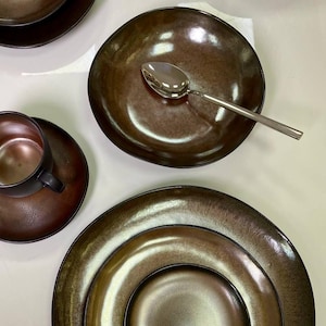 May include: Set of four brown ceramic plates with a metallic sheen. The plates are nested together, with a smaller plate inside a larger plate. The plates have a slightly rippled edge.