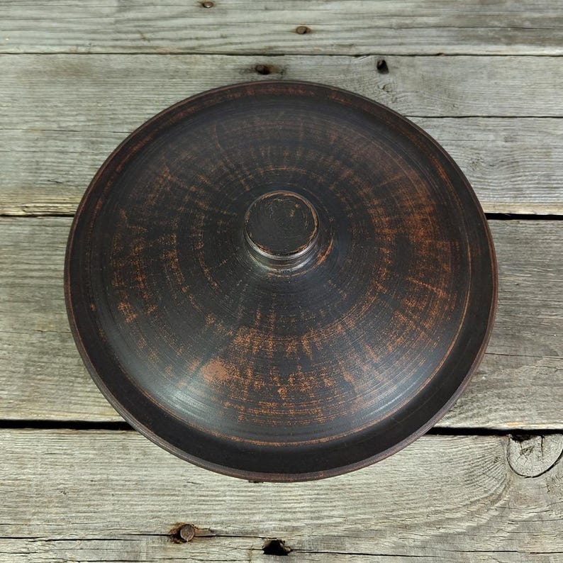 May include: A dark brown ceramic pot lid with a circular knob on top. The lid has a textured surface and a slightly raised rim.