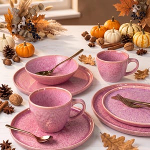 May include: A set of pink ceramic dinnerware featuring speckled glaze, including two mugs, two plates, one bowl, and two gold-toned spoons. The tableware is arranged with autumn-themed decorations like small pumpkins, pinecones, and dried leaves.