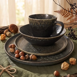May include: A dark grey ceramic dinnerware set consisting of a large round dinner plate, a smaller salad plate, a deep bowl, and a mug. The pieces feature a mottled, reactive glaze finish with lighter grey and brown tones along the edges.