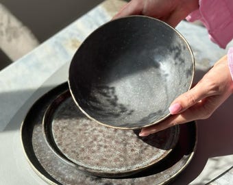 Könnte beinhalten: Ein Set aus drei Keramiktellern in dunkelgrauer Farbe mit einer gesprenkelten Oberfläche. Die Teller sind übereinander gestapelt, wobei der größte Teller unten und der kleinste Teller oben liegt. Die Teller haben einen dezenten Metallic-Glanz.