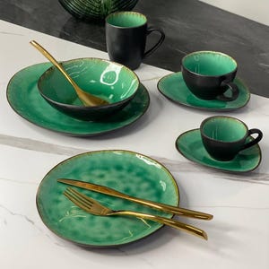 Könnte beinhalten: Ein Set aus grünem und schwarzem Keramikgeschirr mit goldenem Besteck. Das Set enthält einen großen Teller, einen kleineren Teller, eine Schüssel und zwei Teetassen mit Untertassen.