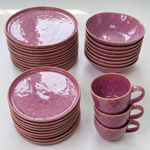 May include: A dinnerware set featuring stacks of round plates, bowls, and three stacked mugs. Each piece has a glossy, mottled magenta and purple glaze with a smooth, reflective surface and a consistent circular shape throughout the collection.