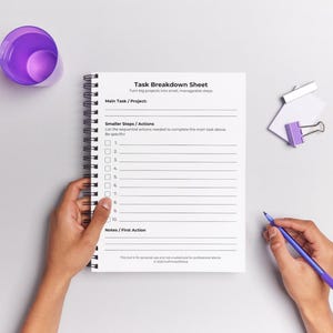 Blank Task Breakdown Sheet shown in a spiral-bound notepad held by two hands against a light grey background. A hand holds a purple pen on the right. A purple cup and a white-and-purple binder clip sit nearby. The worksheet displays the title, subtitle, sections for Main Task/Project, Smaller Steps/Actions with 10 numbered checkbox lines, and Notes/First Action. Footer includes personal use disclaimer and © 2025 ForPrintsOfMind. Soft purple and grey styling props.