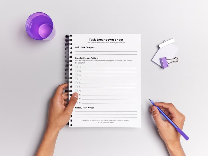 Blank Task Breakdown Sheet shown in a spiral-bound notepad held by two hands against a light grey background. A hand holds a purple pen on the right. A purple cup and a white-and-purple binder clip sit nearby. The worksheet displays the title, subtitle, sections for Main Task/Project, Smaller Steps/Actions with 10 numbered checkbox lines, and Notes/First Action. Footer includes personal use disclaimer and © 2025 ForPrintsOfMind. Soft purple and grey styling props.