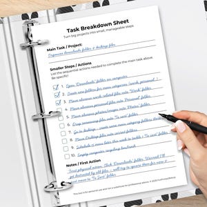 Task Breakdown Sheet printable in an open black-and-white polka dot binder on a wooden desk. A hand holds a pen over the sheet. Filled in with blue handwriting: main task "Organize downloads folder + desktop files" with 10 numbered steps and checkboxes, four checked off. Sections: Main Task/Project, Smaller Steps/Actions, and Notes/First Action. Footer reads "This tool is for personal use and not a substitute for professional advice. © 2025 ForPrintsOfMind."