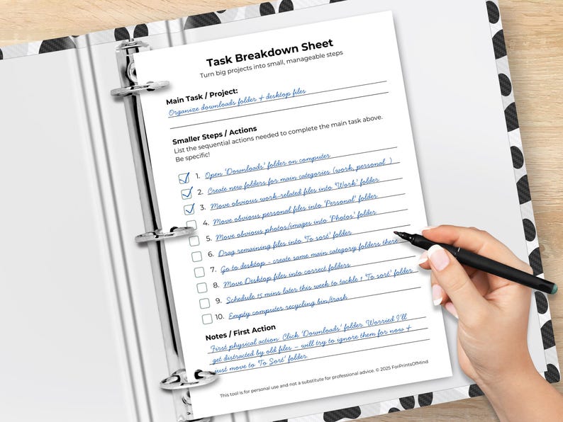 Task Breakdown Sheet printable in an open black-and-white polka dot binder on a wooden desk. A hand holds a pen over the sheet. Filled in with blue handwriting: main task "Organize downloads folder + desktop files" with 10 numbered steps and checkboxes, four checked off. Sections: Main Task/Project, Smaller Steps/Actions, and Notes/First Action. Footer reads "This tool is for personal use and not a substitute for professional advice. © 2025 ForPrintsOfMind."
