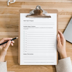 Blank Task Breakdown Sheet on a brown clipboard held by two hands over a wooden desk. A pencil is held in the left hand. A laptop and earbuds are partially visible. The worksheet shows the subtitle "Turn big projects into small, manageable steps," sections for Main Task/Project, Smaller Steps/Actions with 10 numbered checkbox lines, and Notes/First Action. Muted grey-green and white colour palette. Footer includes personal use disclaimer and © 2025 ForPrintsOfMind.