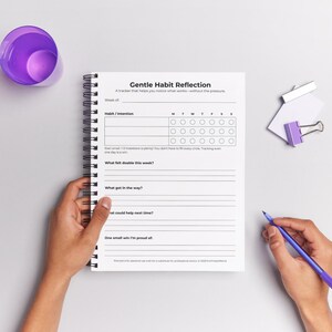 ADHD Habit Tracker printable page shown inside an open spiral-bound notebook, with a hand ready to write on it and a glass of water nearby.