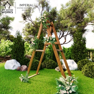 May include: A wooden wedding arch with a triangular A-frame design, decorated with floral arrangements. The arch is set on a green lawn, surrounded by trees and bushes. The text "DIY WOOD WORKS" and "IMPERIAL SYSTEM VERSIONS ARE AVAILABLE" are visible.