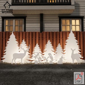 Puede incluir: Arte navideño de madera blanca con siluetas de ciervos, pinos y un belén. El arte está colocado frente a una valla de madera y una casa. El logotipo de DIY Wood Works está en la esquina superior izquierda.