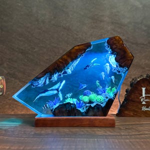 May include: A unique resin art piece depicting an underwater scene with a whale, stingray, and other marine life. The artwork is encased in a clear, angled resin block with a wooden base and edges. The scene is illuminated with a blue light.