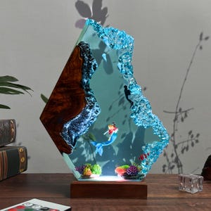 Pode incluir: Escultura decorativa em resina com uma sereia, um mergulhador e um recife de coral. A escultura tem uma base de madeira e um corpo de resina transparente tingido de azul com uma borda texturizada e cristalina. A sereia tem cabelo ruivo e uma cauda azul.
