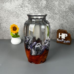 May include: A clear glass vase with a distinctive design. The base has a dark wood grain pattern, leading to a rocky, purple and grey section. The top is clear, and the vase is cylindrical. A small, crocheted sunflower is in the background.