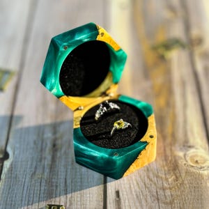 Può includere: Un portagioie esagonale aperto, con esterno in resina verde e gialla. Contiene due anelli su un interno in velluto nero. La scatola è in legno e resina, appoggiata su una superficie di legno.