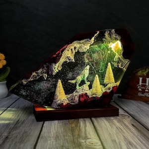 Può includere: Lampada decorativa con una statuina di lupo che ulula in un paesaggio in resina. La lampada ha un esterno rosso scuro e oro e una base in legno. La scena interna comprende alberi in miniatura e un cielo stellato, illuminato con luce verde e gialla.