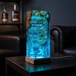 May include: A rectangular table lamp featuring a block of clear resin and natural wood. The resin contains a textured, moss-covered rock formation illuminated from below with a bright blue light, creating a swirling, ethereal glow effect.