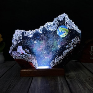 May include: A space-themed night light with a clear, textured exterior. Inside, a colourful galaxy scene is visible, with an astronaut figure and a miniature Earth. The light sits on a wooden base, creating a unique decorative piece.