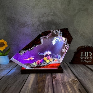 May include: A unique resin art piece featuring a vibrant underwater scene with axolotls and colorful coral. The artwork is encased in a geometric shape with a wooden base, illuminated by internal lighting. The piece is a decorative accent.