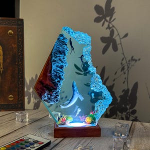 May include: A decorative resin lamp depicting an underwater scene with a whale, divers, and coral. The lamp has a wooden base and a blue, textured edge, with a remote control for the LED lights. The lamp is approximately 25 cm tall.