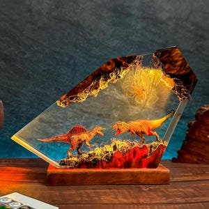 May include: A decorative lamp featuring two dinosaur figures encased in clear resin. The dinosaurs are orange and red, set against a backdrop of amber and brown resin. The lamp sits on a wooden base.