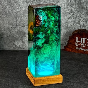 May include: A rectangular resin lamp with a wooden base. The lamp features a vibrant display of green and blue hues, with a hint of yellow. The base is a natural wood color. The lamp is a decorative lighting fixture.
