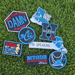 May include: A collection of embroidered patches featuring various designs, including a blue and white sneaker, a black and white 'La' logo, a black patch with the text 'Mind Your Own Damn Business', a white patch with the text 'I'm Speaking', a black patch with the text 'Yes We Can', a black patch with the text 'Harris 47', a black patch with the text 'The First But Not The Last Kamala Harris', a black patch with the text 'MYODB Mind Your Own Damn Business -Walz', and a black patch with a blue sneaker and a string of pearls.