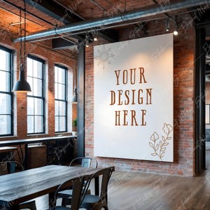 Op de afbeelding: Een witte canvas mockup met de tekst "YOUR DESIGN HERE" in bruine letters en een eenvoudig bloemenontwerp in de rechterbenedenhoek. Het canvas hangt aan een bakstenen muur in een loft-achtige ruimte met een houten vloer en een tafel met stoelen.