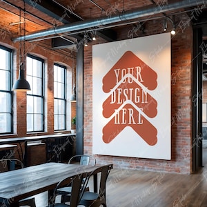 Op de afbeelding: Een wit canvas met de tekst "YOUR DESIGN HERE" in rood-oranje, gemonteerd op een bakstenen muur in een loft-achtige ruimte met een houten vloer en een tafel met stoelen.