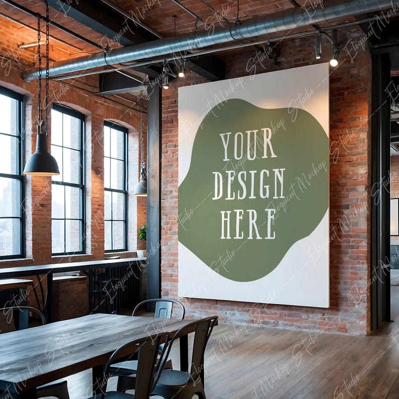Op de afbeelding: Een witte canvas mockup met een groene abstracte vorm en de tekst "YOUR DESIGN HERE" in een rustieke loft-setting met blootliggende bakstenen muren, een houten vloer en een tafel met stoelen.