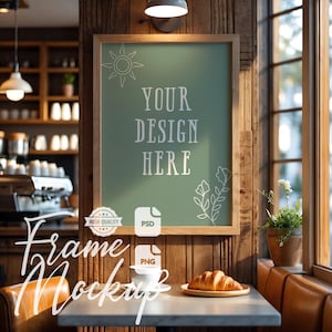 以下が含まれることがあります： 緑色の背景に「YOUR DESIGN HERE」というテキストが入った額入りのモックアップ。 フレームは木の壁に掛けられています。 クロワッサン、コーヒーカップ、植物のあるテーブルがフレームの前にあります。