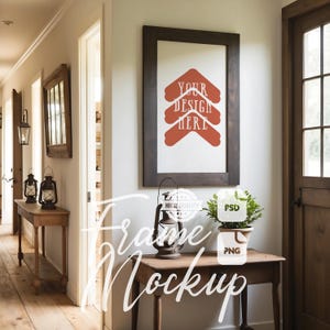 Puede incluir: Un marco de madera marrón con un paspartú blanco y un gráfico rojo que dice "Your Design Here" colgado en una pared blanca en un pasillo con suelo de madera. Hay una mesa pequeña con una linterna y una planta encima.