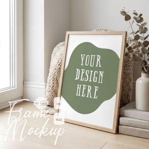 May include: A wooden framed mockup with the text "YOUR DESIGN HERE" on a green abstract shape. The frame is leaning against a beige knitted throw. A vase with greenery and a stack of books are visible.