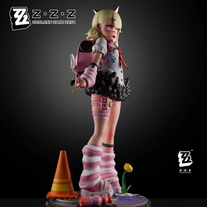 Puede incluir: Una figura de anime rosa y negra de una niña con cuernos, que lleva una camisa a rayas rosa y blancas, una falda negra y calcetines a rayas rosa y blancas. La figura está de pie sobre una base gris con un cono de tráfico negro y naranja.