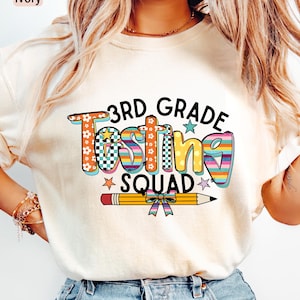 May include: Ivory-colored t-shirt with a colorful graphic that reads "3rd Grade Testing Squad." The text features floral, checkered, and striped patterns. A yellow pencil with a bow is below the text. The shirt is a casual style.