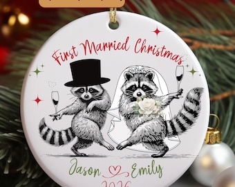 May include: A round white ceramic ornament featuring two raccoons dressed as a bride and groom. The text reads First Married Christmas, Jason, Emily, and 2026. The groom wears a top hat, and the bride wears a veil and holds a white flower.