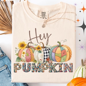 May include: Ivory-coloured t-shirt featuring the text "Hey Pumpkin" and a graphic of three pumpkins with floral and patchwork designs. The shirt is folded next to a pair of blue jeans and several pumpkins.