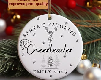 May include: White ceramic Christmas ornament with a gold string. The ornament features the text "SANTA'S FAVORITE Cheerleader" with a cheerleader graphic, snowflakes, and the name "EMILY 2025".