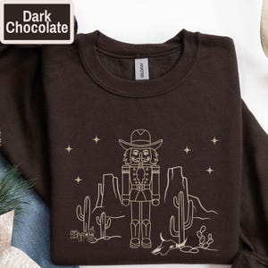 May include: Dark chocolate-colored sweatshirt featuring a beige outline of a cowboy nutcracker in a desert scene with cacti, stars, and a cow skull. The text "Dark Chocolate" is in a white box in the upper left corner.