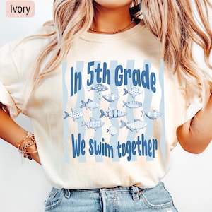May include: An ivory short-sleeved t-shirt featuring a blue graphic print of various fish and the text In 5th Grade We swim together. The design includes light blue vertical wavy stripes behind the fish and blue lettering.