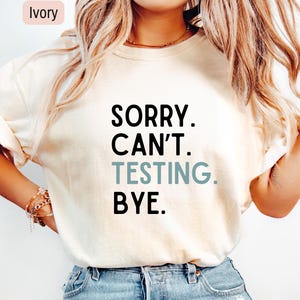 May include: Ivory-colored t-shirt with the text "SORRY. CAN'T. TESTING. BYE." in black and teal lettering. The shirt is a casual style, suitable for everyday wear.