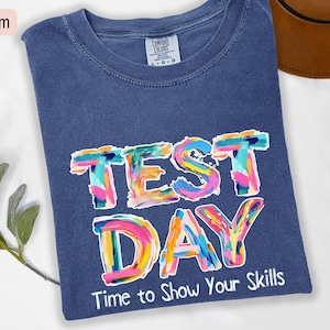 May include: A denim blue t-shirt featuring the words "TEST DAY" in vibrant, artistic lettering. The text "Time to Show Your Skills" is printed below. The shirt is a Comfort Colors brand.