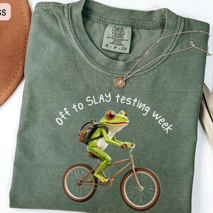 May include: Moss green t-shirt with a frog riding a bicycle graphic. The frog has a backpack and red eyes. The text "off to SLAY testing week" is arched above the frog. A silver necklace with a pendant is on the shirt.