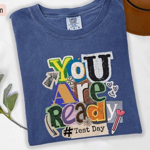 May include: Denim blue t-shirt with the words "You Are Ready" in a collage style. The design includes a pencil, hearts, and the hashtag "Test Day". The shirt is a Comfort Colors brand.