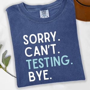 May include: A denim blue t-shirt with the words "SORRY. CAN'T. TESTING. BYE." in white and teal lettering. The shirt is folded and has a crew neck. A brown hat is partially visible in the background.